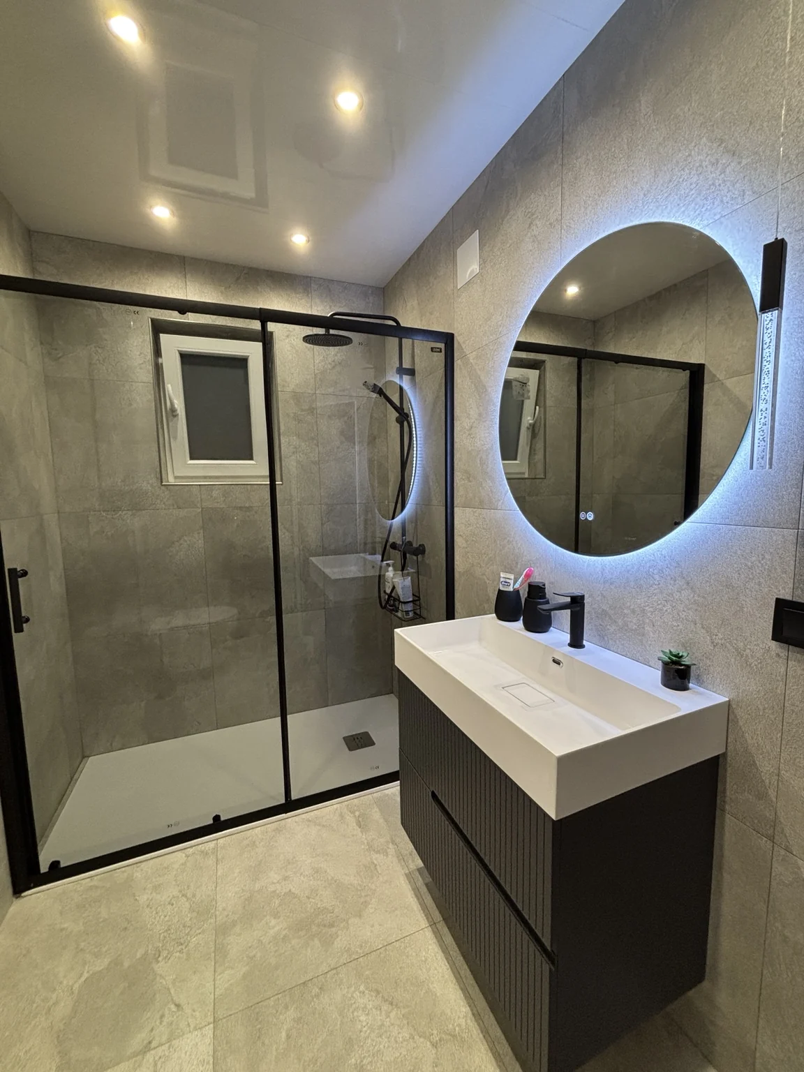 Baño reformado con ducha, mampara negra y espejo circular iluminado