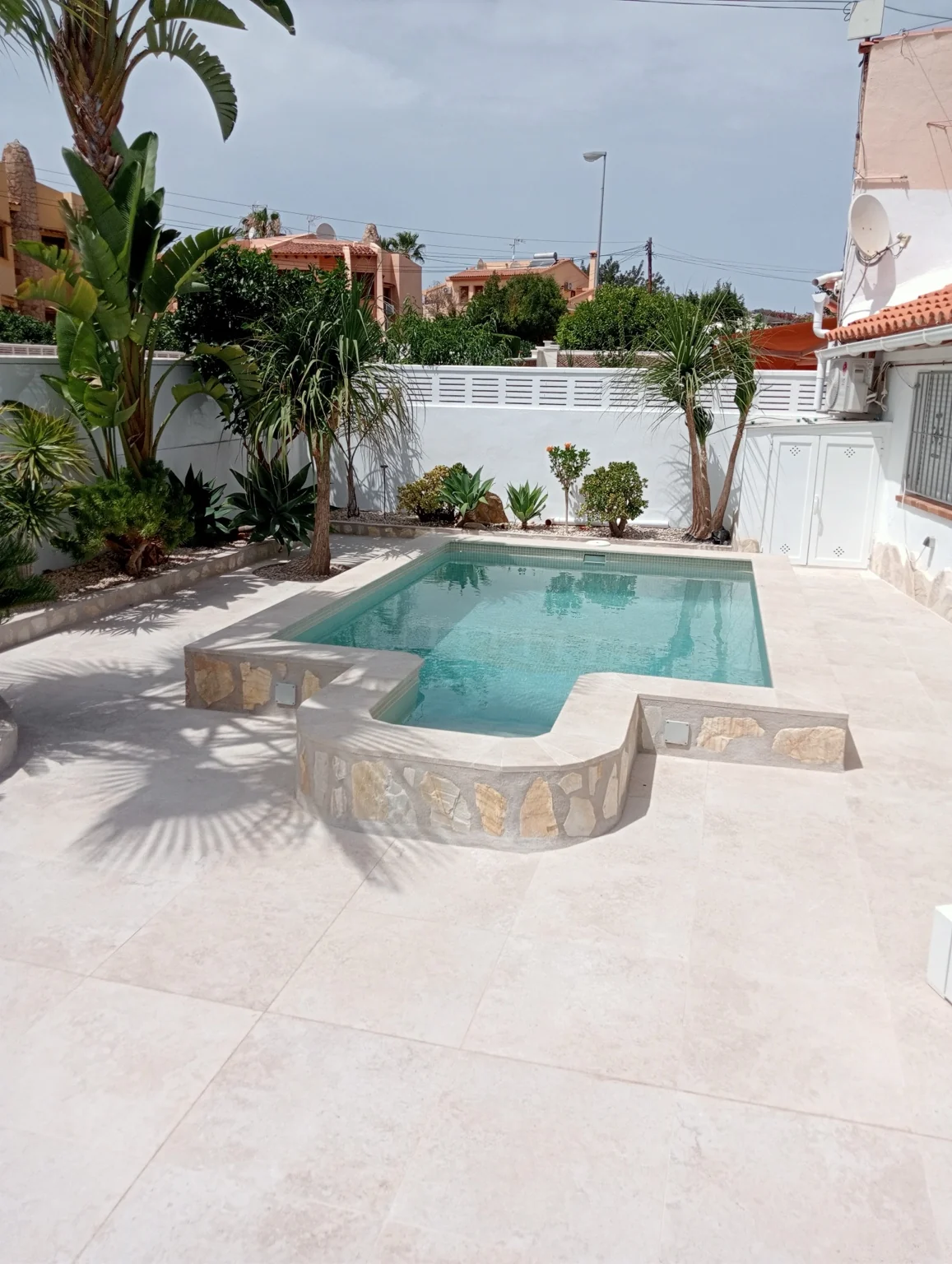 Piscina exterior con revestimiento de piedra, pavimento claro y jardín